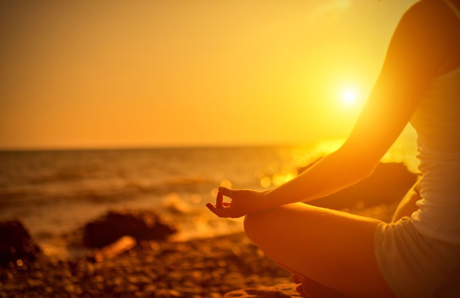 Silhouette d’une personne assise en tailleur au bord de la mer au soleil couchant, la main posée sur le genou en jnana mudra.